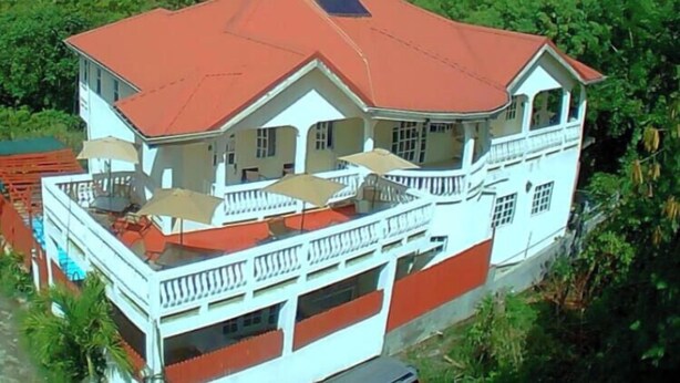 A large, two-story building with a red roof and white walls, surrounded by lush greenery and mountains in the background.