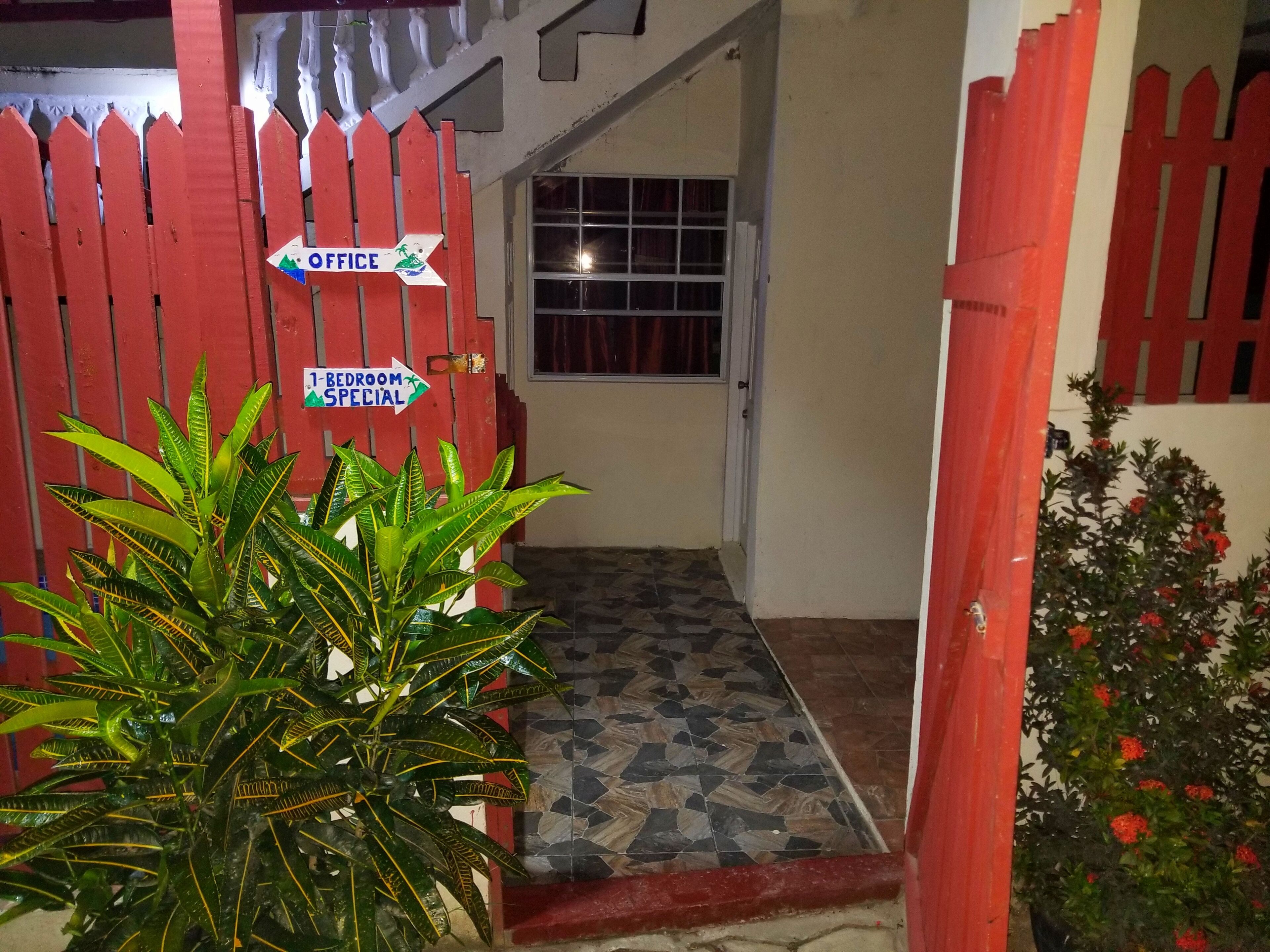Un couloir avec une petite clôture en bois peinte en rouge, un pot de fleurs et des panneaux indiquant un bureau et une chambre d’amis.
