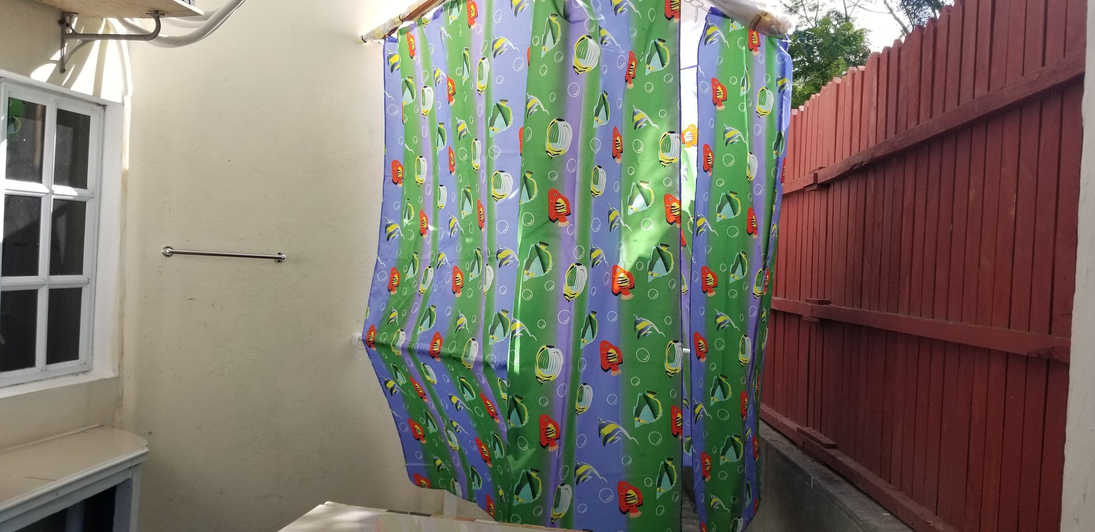 A bathroom with a shower curtain featuring a fish pattern, a window, and a red fence.