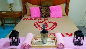 A bed with a 'HAPPY' message and a heart made of rose petals, a bottle of champagne, two glasses, and two lanterns on a tray.