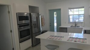 A modern kitchen with stainless steel appliances, a white island with bar stools, and a window providing natural light. - Walk to the beach! Clean and new, remodeled  cozy 3 bedroom 2 bath cottage house (Venice)
