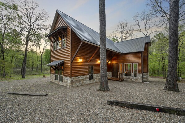A log cabin with a stone foundation, large windows, and a gravel driveway.