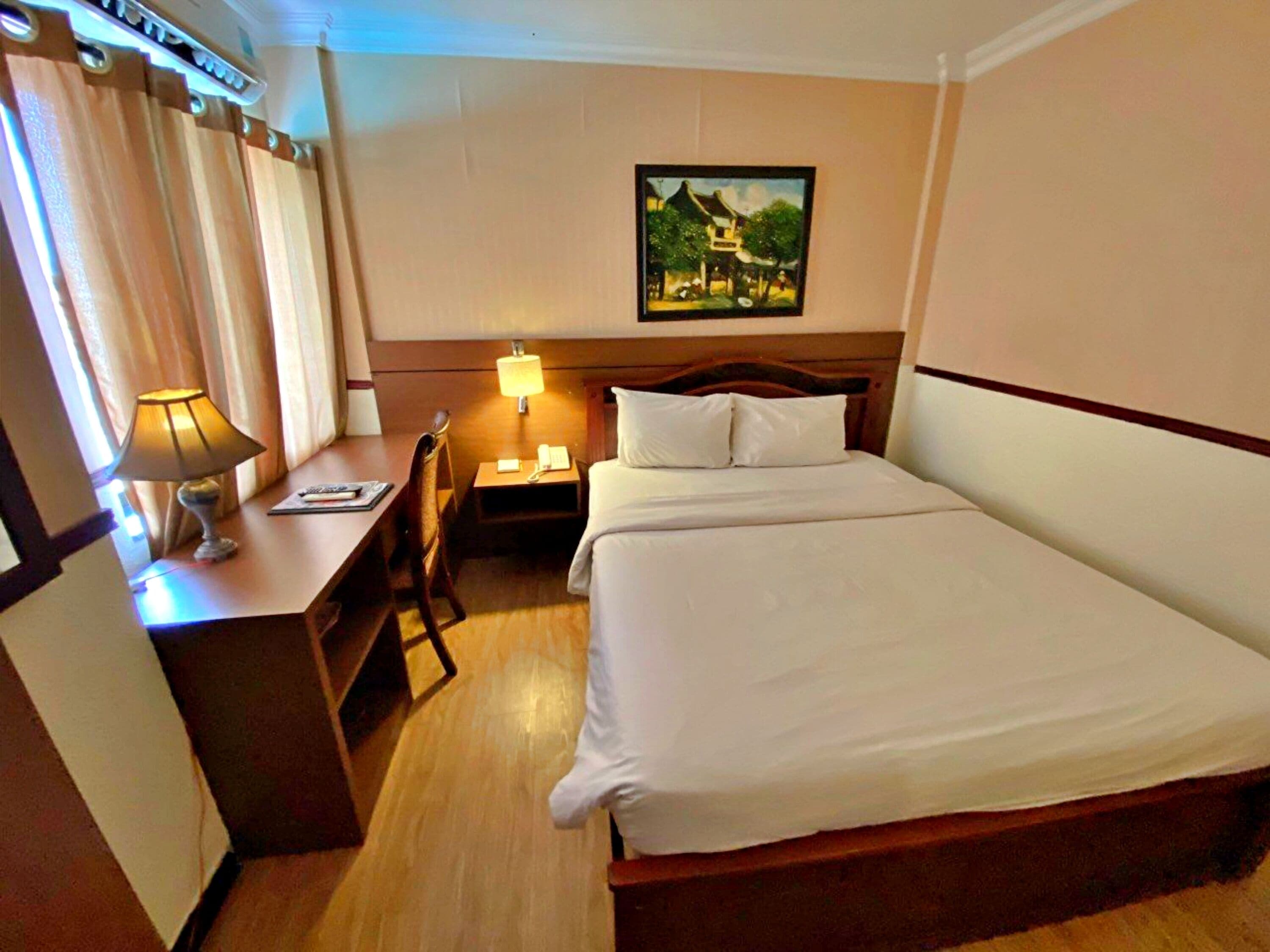 A hotel room with a bed, a desk with a lamp, a chair, and a painting on the wall.