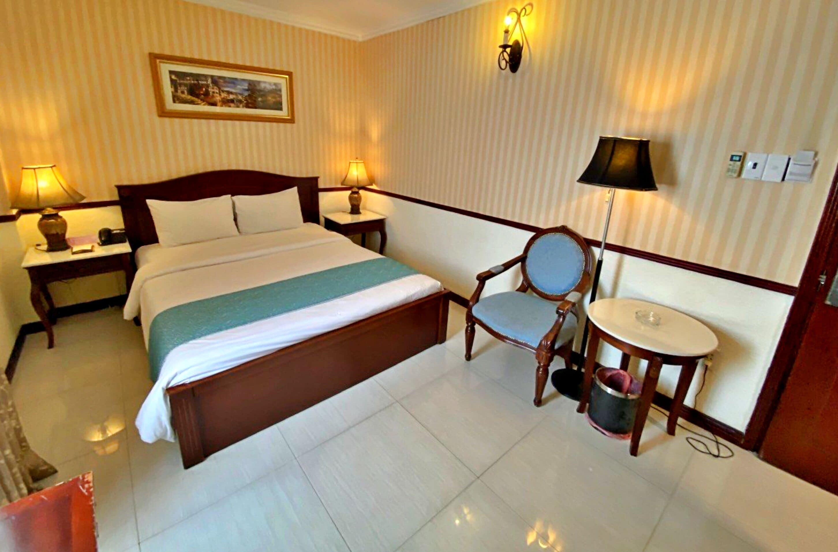 A hotel room with a large bed, two bedside tables with lamps, a chair, a small round table, and a wall-mounted light fixture.