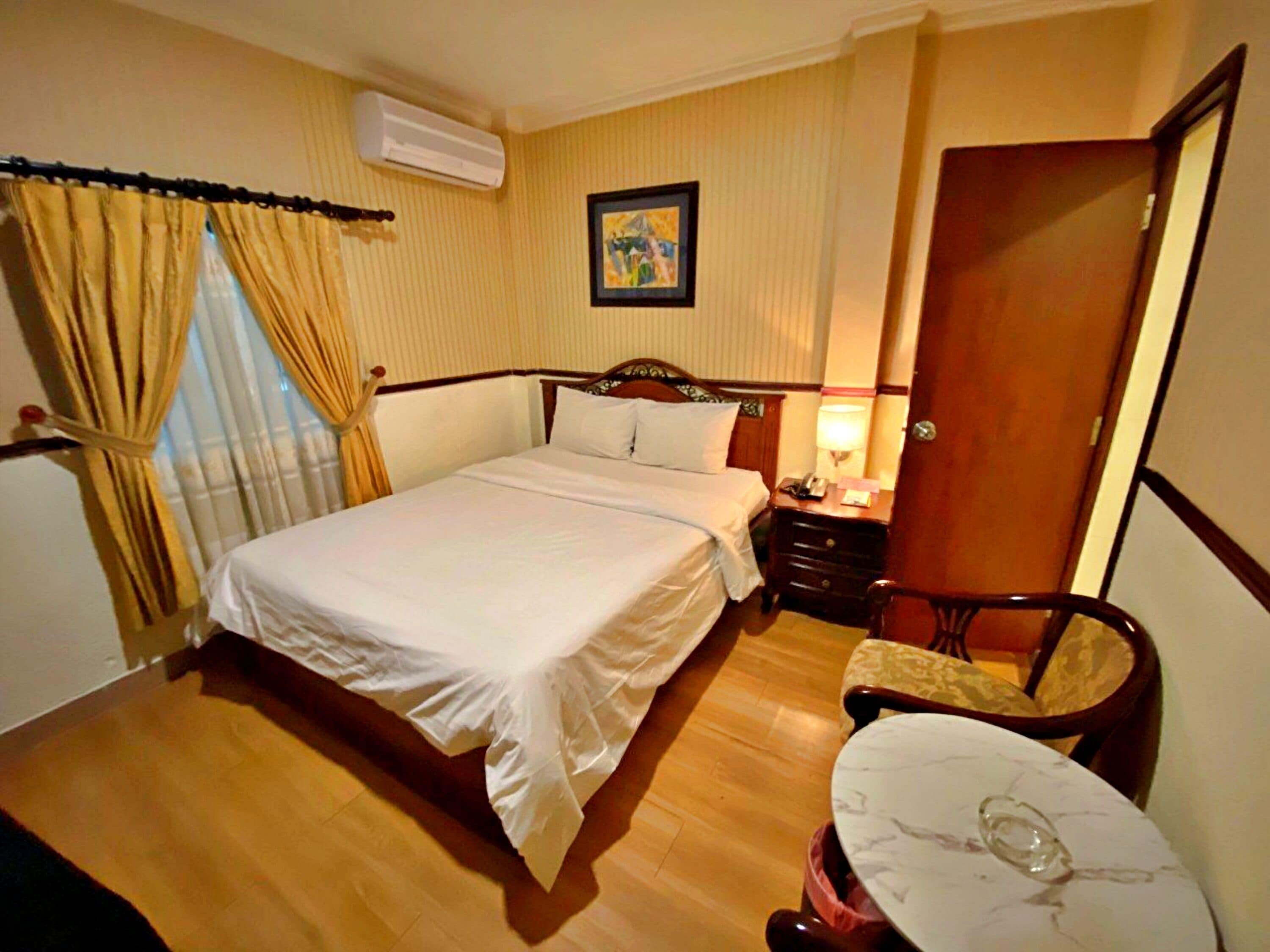 A hotel room with a bed, bedside table, lamp, chair, and a small table with a glass.