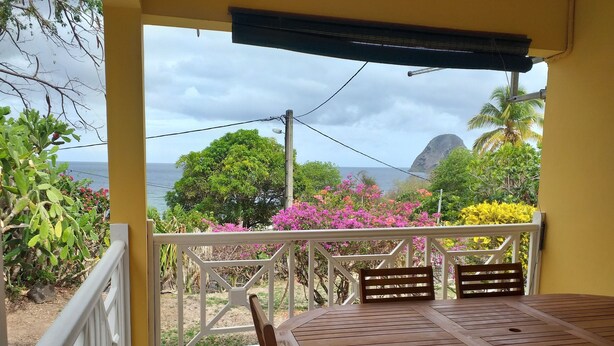 A covered outdoor seating area with a table and chairs, overlooking a scenic view of the ocean and lush vegetation. - Les Océanides Villa 2 with sea view (Le Diamant)