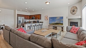 A modern living room with a sofa set, a dining area with bar stools, a kitchen with stainless steel appliances, and a fireplace. - 3BR w fireplace, gaming TVs, mountain views! (Bellemont)