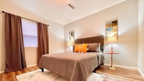 Quarto com cama, cortinas, mesa de cabeceira, abajur e um quadro na parede.