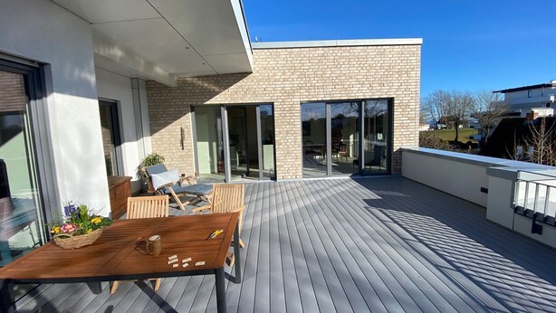 Une terrasse extérieure moderne, avec une table et des chaises en bois, un panier de fleurs et une porte-fenêtre coulissante donnant sur un patio abrité.
