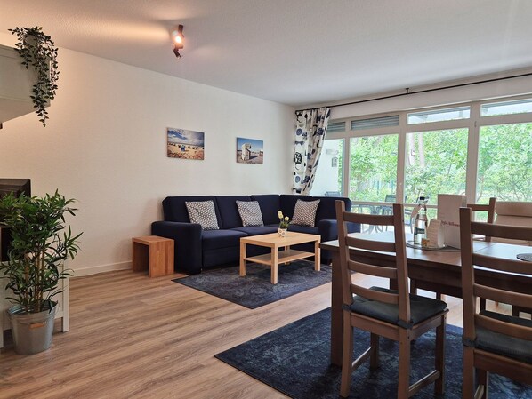A modern living room with a sofa, coffee table, and dining area. There are two paintings on the wall, a window with curtains, and a plant on the left.