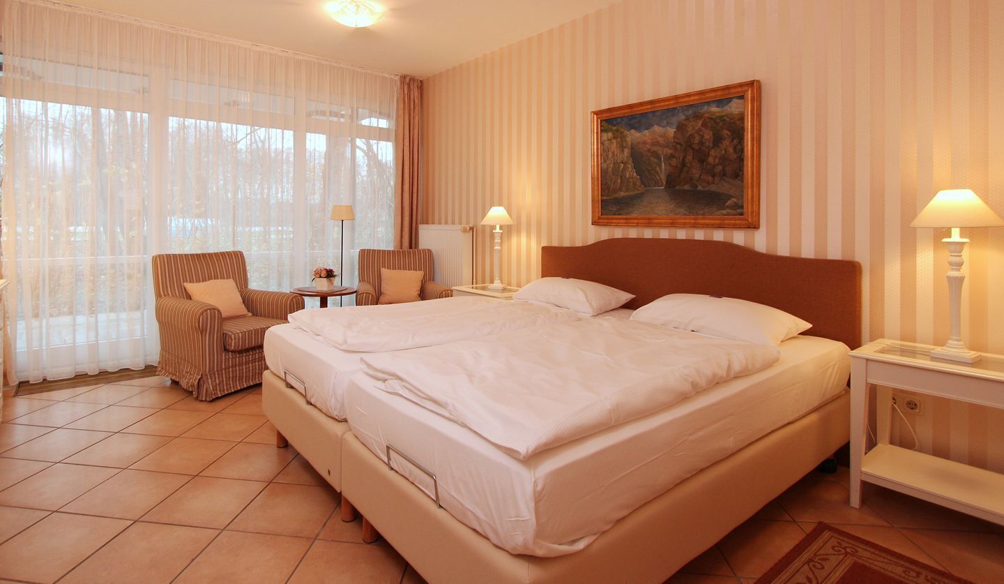 A hotel room with a large bed, two bedside tables, a chair, a painting on the wall, and a window with sheer curtains.