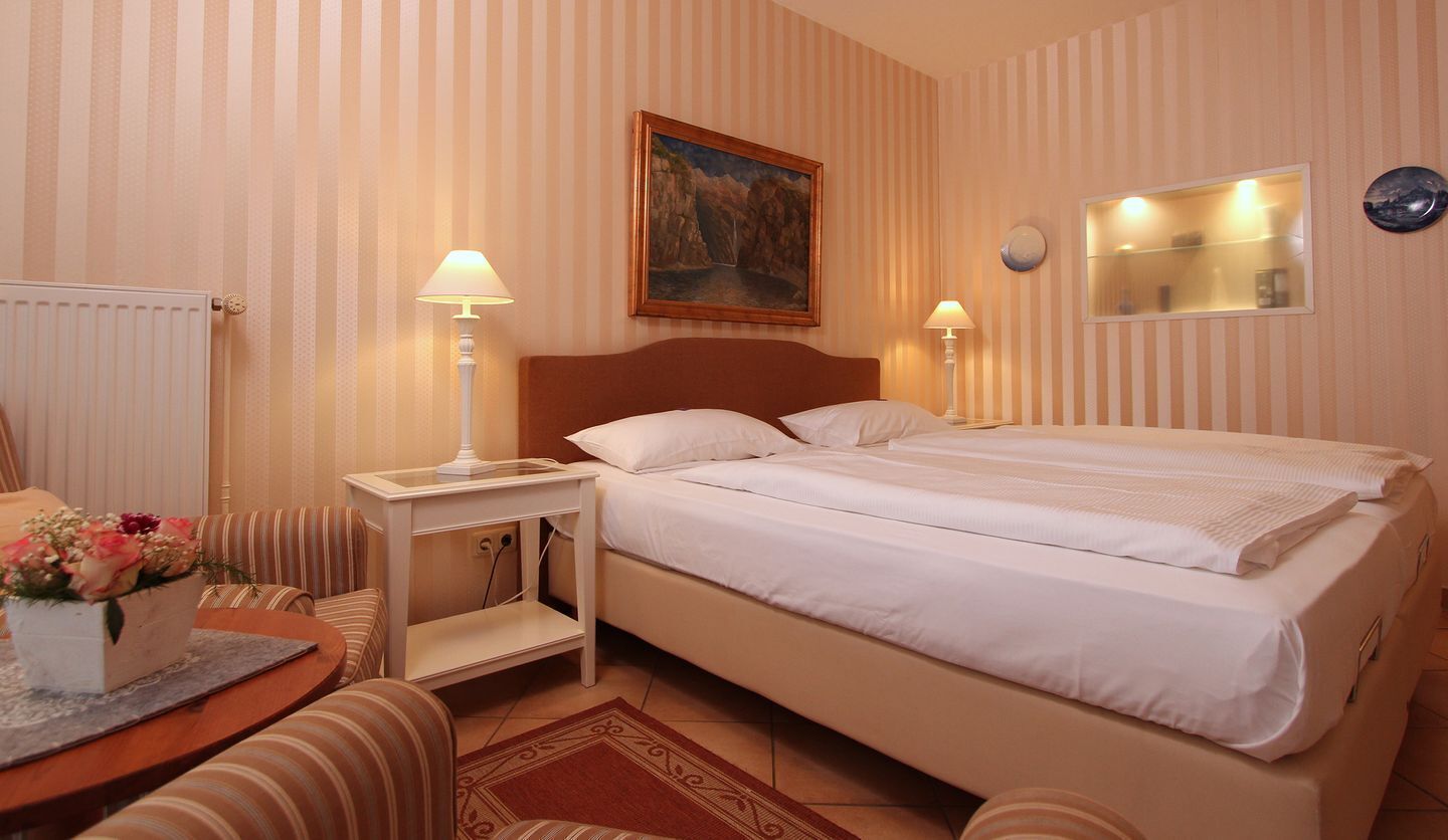 A hotel room with a bed, two bedside tables with lamps, a painting on the wall, and a small table with flowers.