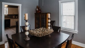 Una mesa de comedor con un centro de mesa, rodeada de sillas, en una habitación con una ventana y un aparador.