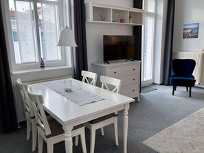 Ett rum med ett vitt bord och stolar, en tv på ett vitt skåp, en blå stol och ett fönster med gardiner.