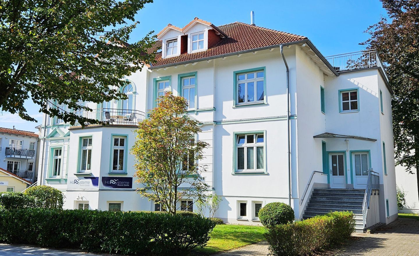 A multi-story building with a white facade and green trim, featuring a staircase leading to the entrance.