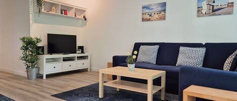 A modern living room with a dark blue sofa, a wooden coffee table, a flat-screen TV on a white cabinet, and a small plant.