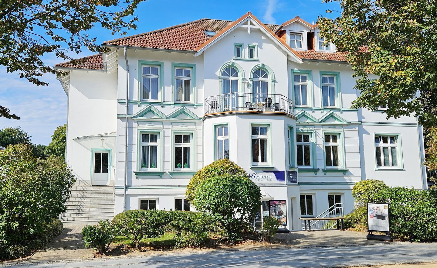 Ein großes, weißes Gebäude mit grünen Akzenten und zahlreichen Fenstern, mit einem Balkon und einer Treppe zum Eingang.