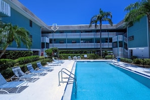 A pool area with lounge chairs and a clear blue pool, surrounded by a multi-story building with balconies.
