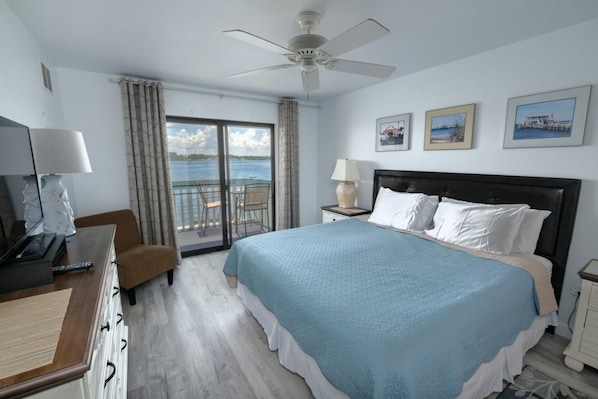 A hotel room with a large bed, a ceiling fan, a desk with a lamp, a chair, and a view of the sea through a sliding glass door.