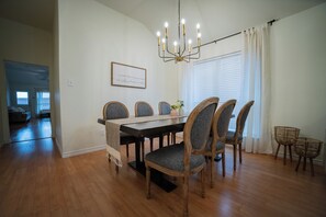 A dining area with a wooden table, grey upholstered chairs, a chandelier, and a window with blinds.