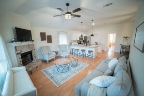 A spacious living room with a fireplace, a ceiling fan, and a kitchen in the background.