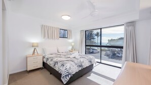 Un dormitorio con cama, mesitas de noche, ventilador de techo, ventana con cortinas y vistas al exterior.