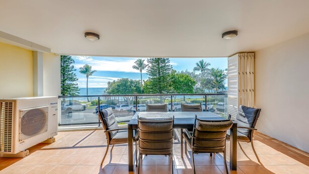Un balcón con mesa y sillas, vistas al mar y palmeras, y un aire acondicionado Panasonic.