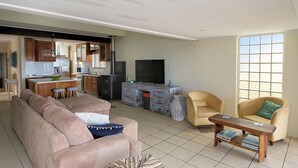 Una sala de estar con sofá, sillones, una mesita de centro, televisor y una zona de cocina con armarios y estufa.