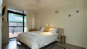 Un dormitorio con cama, una silla, un taburete, un ventilador de techo y vistas al exterior a través de una ventana con cortina.