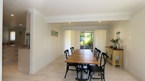 A spacious dining area with a long wooden table and black chairs, leading to a kitchen and a view of greenery outside.