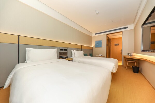 A hotel room with two beds, a desk, a chair, a trash bin, and a door leading to another room.
