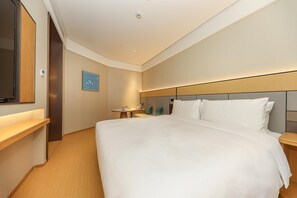 A hotel room with a large bed, a bedside table, a small table with a lamp, a TV mounted on the wall, and a framed picture on the wall.