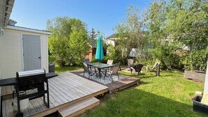 A backyard with a wooden deck, a grill, a table with chairs, and a green umbrella. - Hardy’s cute and cozy beach cottage  (Pointe-du-Chene)
