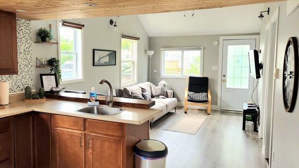 A kitchen with wooden cabinets, a sink, and a countertop. There is a dining area with a sofa and a chair. - Hardy’s cute and cozy beach cottage  (Pointe-du-Chene)