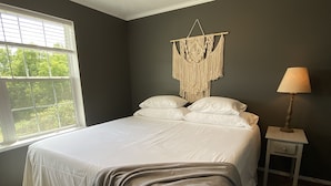 A bedroom with a bed, a bedside table with a lamp, a macrame wall hanging, and a window with blinds. - Natures Crest Retreat, Hocking Hills (Logan)