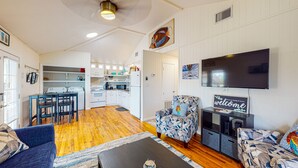 A modern living room with a sofa, armchair, and a coffee table. There is a kitchen area with white cabinets and a flat-screen TV mounted on the wall. - In Town, Pet Friendly, Golf Cart Accessible (Port Aransas)
