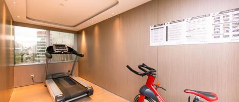 A gym with a treadmill and a stationary bike, a window with a city view, and a wall with informational posters.