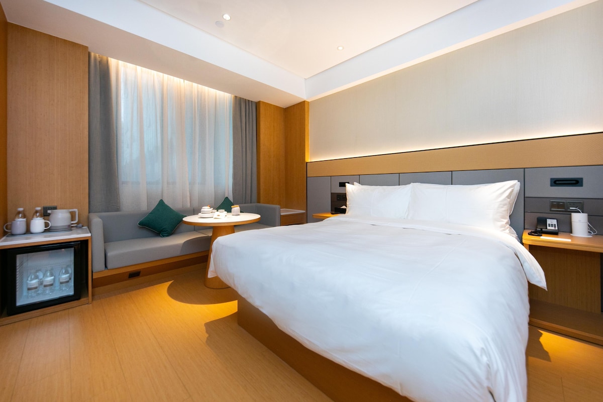 A modern hotel room with a large bed, a sofa, a small round table, and a minibar.