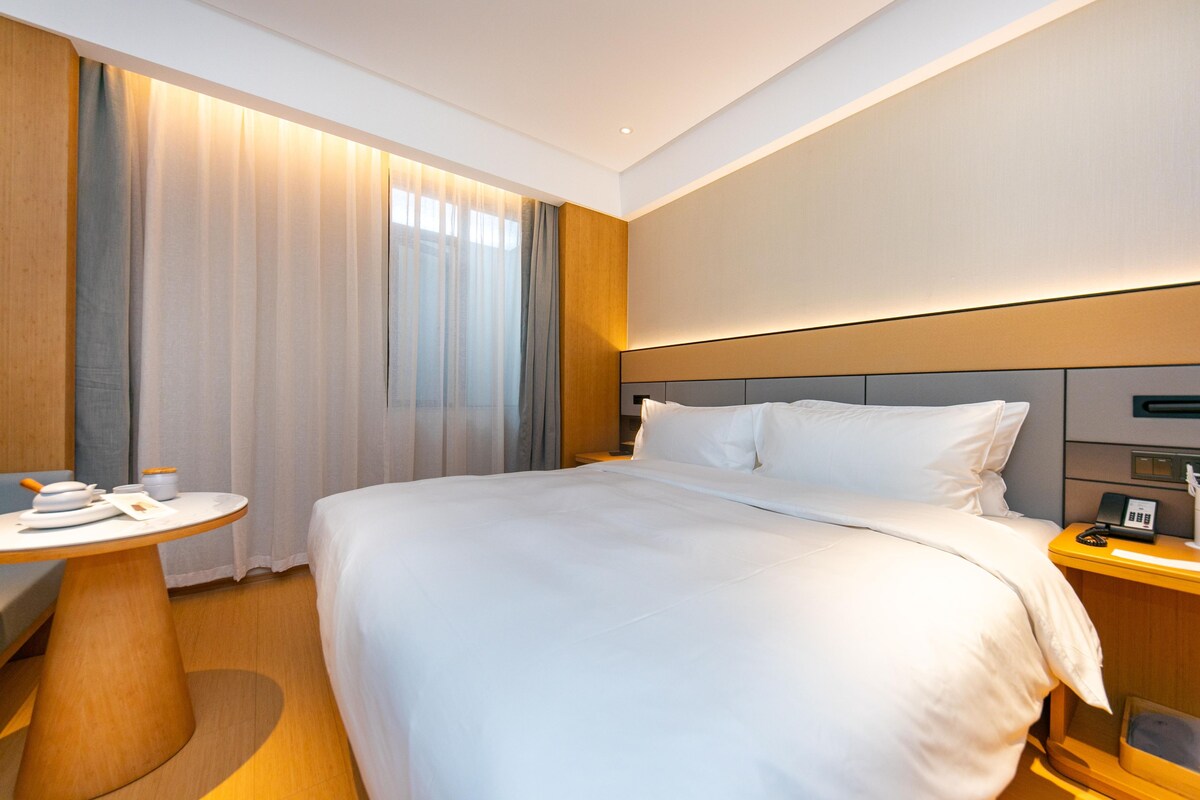 A hotel room with a large bed, a bedside table, a lamp, and a window with curtains.