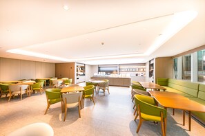 A modern dining area with tables and chairs, a kitchen in the background, and a large window on the right.