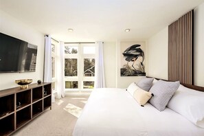 A modern bedroom with a large bed, a flat-screen TV, a wooden console, and a view of the outdoors through a glass door.