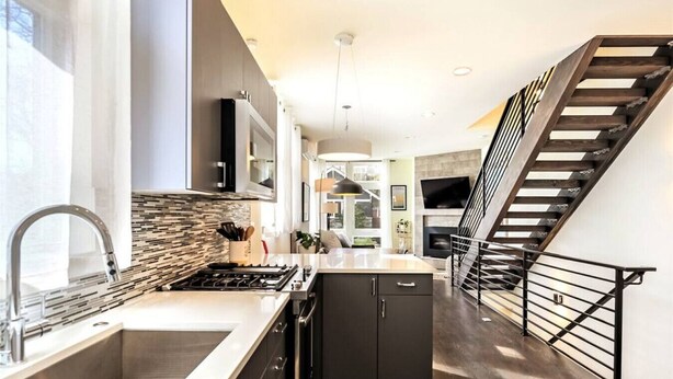 A modern kitchen with a central island, stainless steel appliances, and a black staircase with a metal railing. - Modern 3 Bedroom in Queen Anne with Rooftop + AC (Seattle)