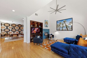 A modern living room with a fireplace, a dining area, and a large TV. - Designer Home-like Condo, Pools, Dog Park (PALM SPRINGS)
