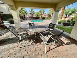 A covered patio with a round table and chairs, a pool in the background, and palm trees.