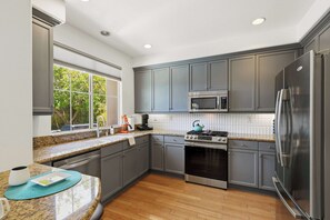 A modern kitchen with grey cabinets, stainless steel appliances, a window with a view of greenery, and a granite countertop. - Designer Home-like Condo, Pools, Dog Park (PALM SPRINGS)