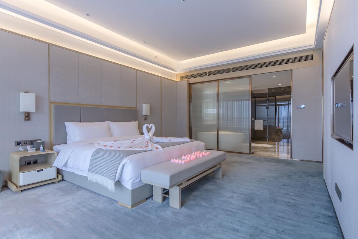 Een moderne hotelkamer met een groot bed, nachtkastjes, een bankje en een glazen schuifdeur naar een balkon.