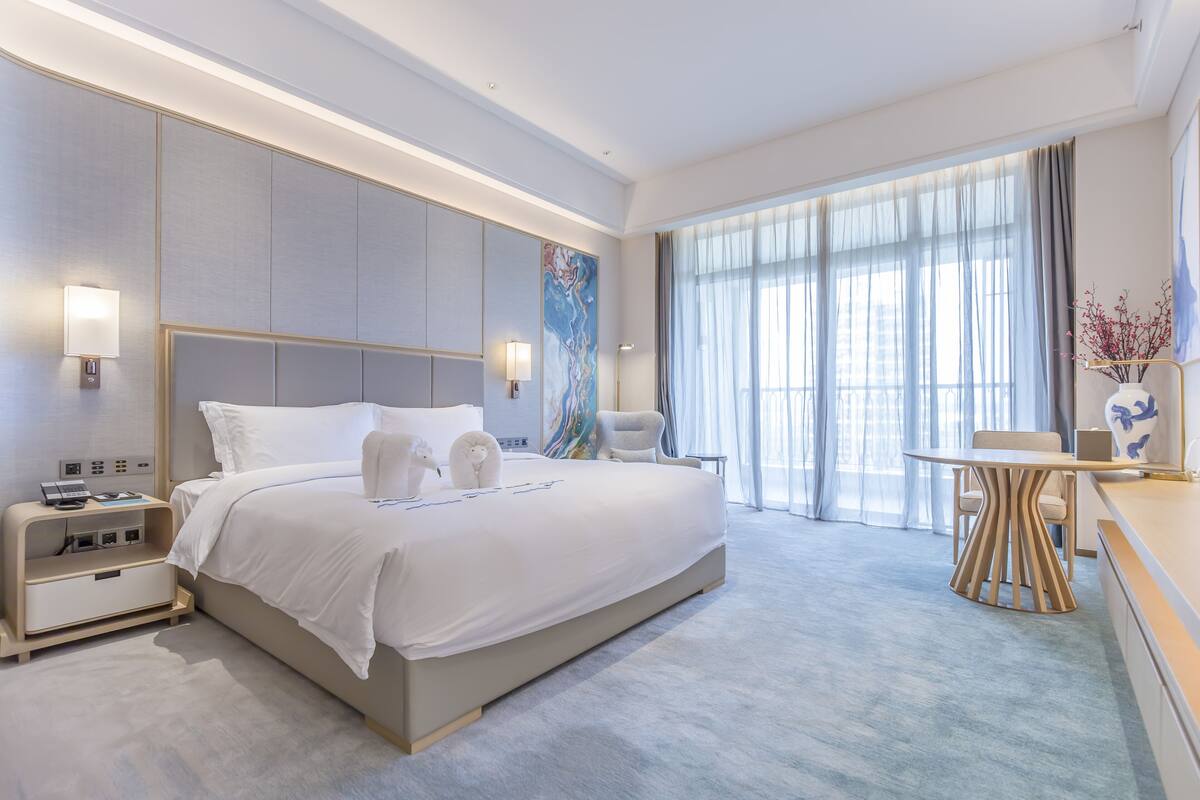 Een moderne hotelkamer met een groot bed, nachtkastjes, een bureau en uitzicht op de stad door grote ramen.