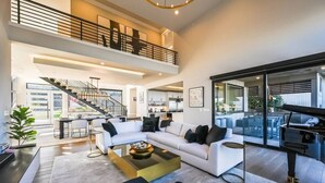 A spacious living area with a white sofa, black and white cushions, a coffee table, and a piano. A staircase leads to an upper level with a balcony.
