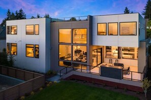 Ein modernes, zweistöckiges Haus mit großen Fenstern, einem Balkon und einem sichtbaren Innenraum mit Möbeln.