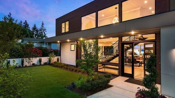 A modern two-story house with a well-maintained lawn, a visible staircase leading to the upper floor, and a large glass door entrance.